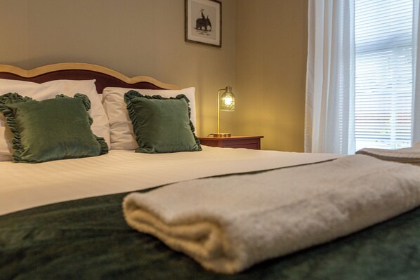 A neatly made bed with a green blanket and pillows, a bedside lamp, and a framed picture on the wall.