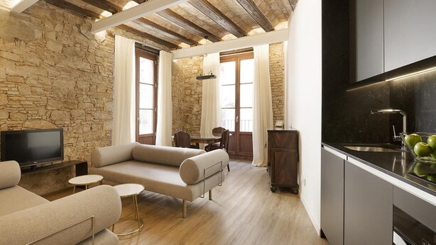 A modern living room with a stone wall, wooden ceiling beams, a sofa, a TV, and a kitchen area with a sink and apples.