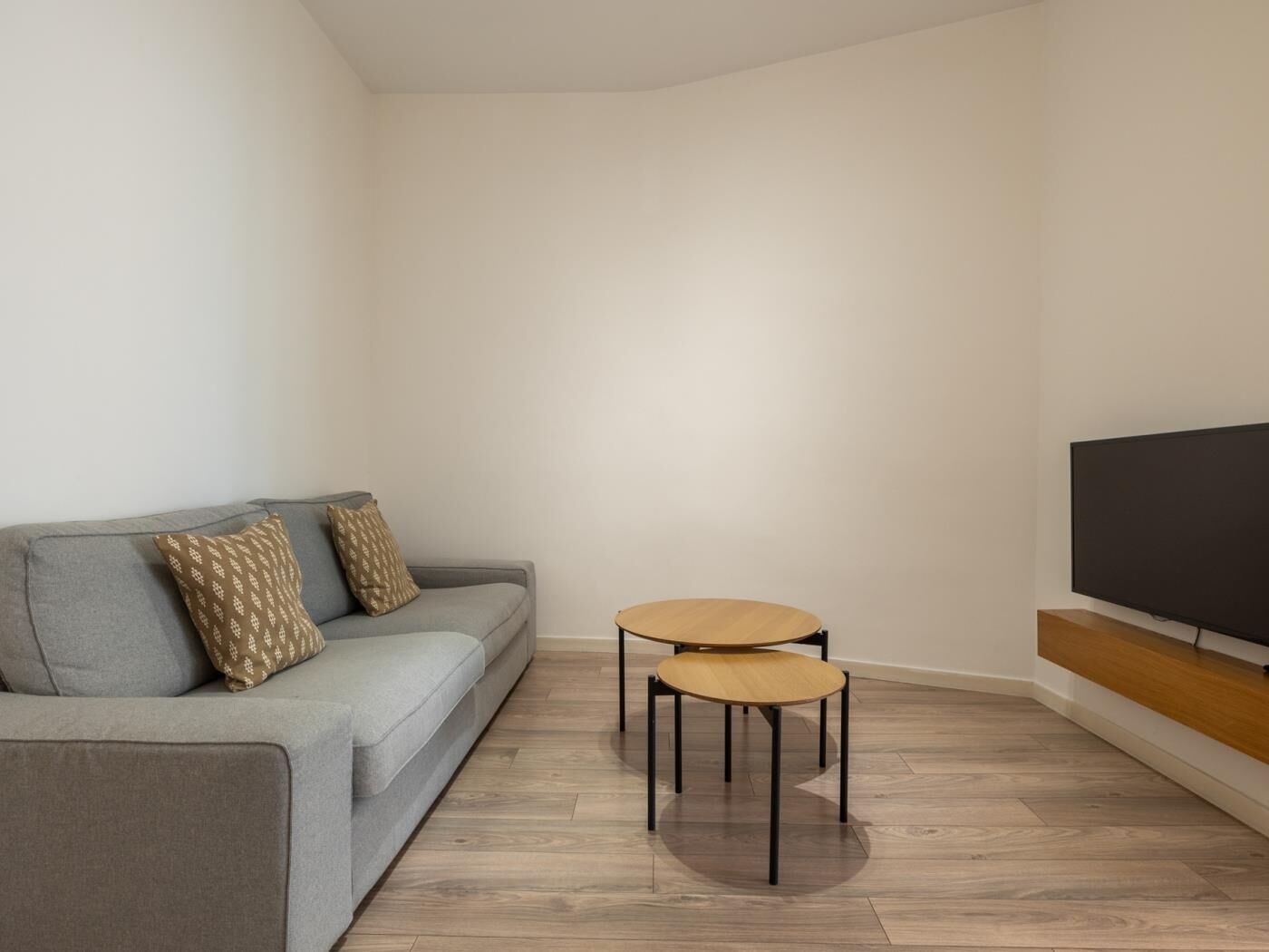 A modern living room with a grey sofa, two round wooden tables, and a flat-screen TV mounted on the wall.