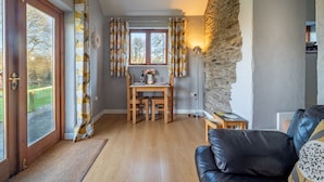 A cozy living room with a black leather sofa, a dining table with chairs, a stone wall, and a view through a large window. - Fantastic couples escape to the country. Quiet, rural location, pet friendly. (Boncath)