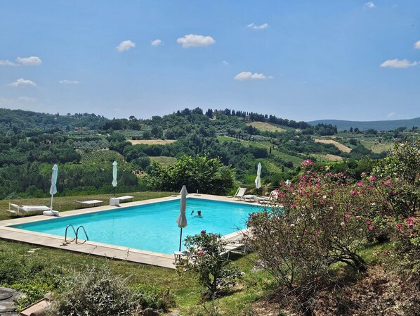 A pool with lounge chairs and umbrellas, surrounded by lush greenery and hills in the background.