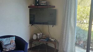 A living room with a flat-screen TV mounted on the wall, a blue sofa with a decorative pillow, a small table with a basket and books, and a window with a curtain. - 2 bedrooms, large pool, stunning views of San Gimignano, Tuscany, private garden (San Gimignano, Tuscany)
