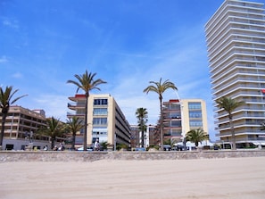 Een hotel aan het strand met meerdere verdiepingen, palmbomen en een helderblauwe lucht.