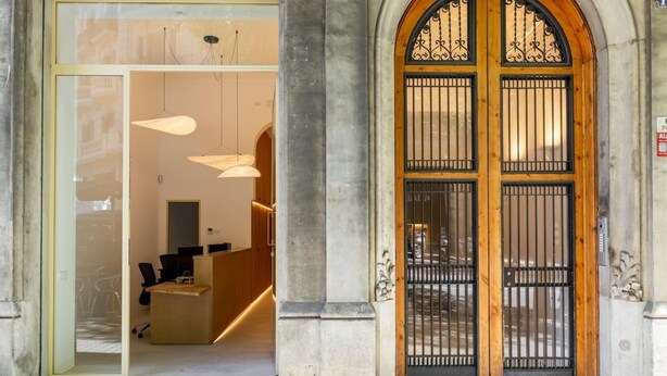A building entrance with a glass door leading to a reception area, a wooden door with metal bars, and a stone facade. - DELUXE THREE BEDROOM APARTMENT (Barcelona)