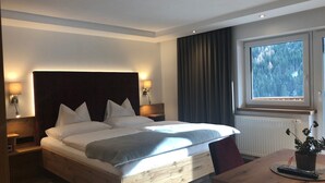 A modern hotel room with a large bed, wooden headboard, two bedside lamps, a window with a forest view, and a wooden table with a remote control. - Hotel Garni Central (Kappl)
