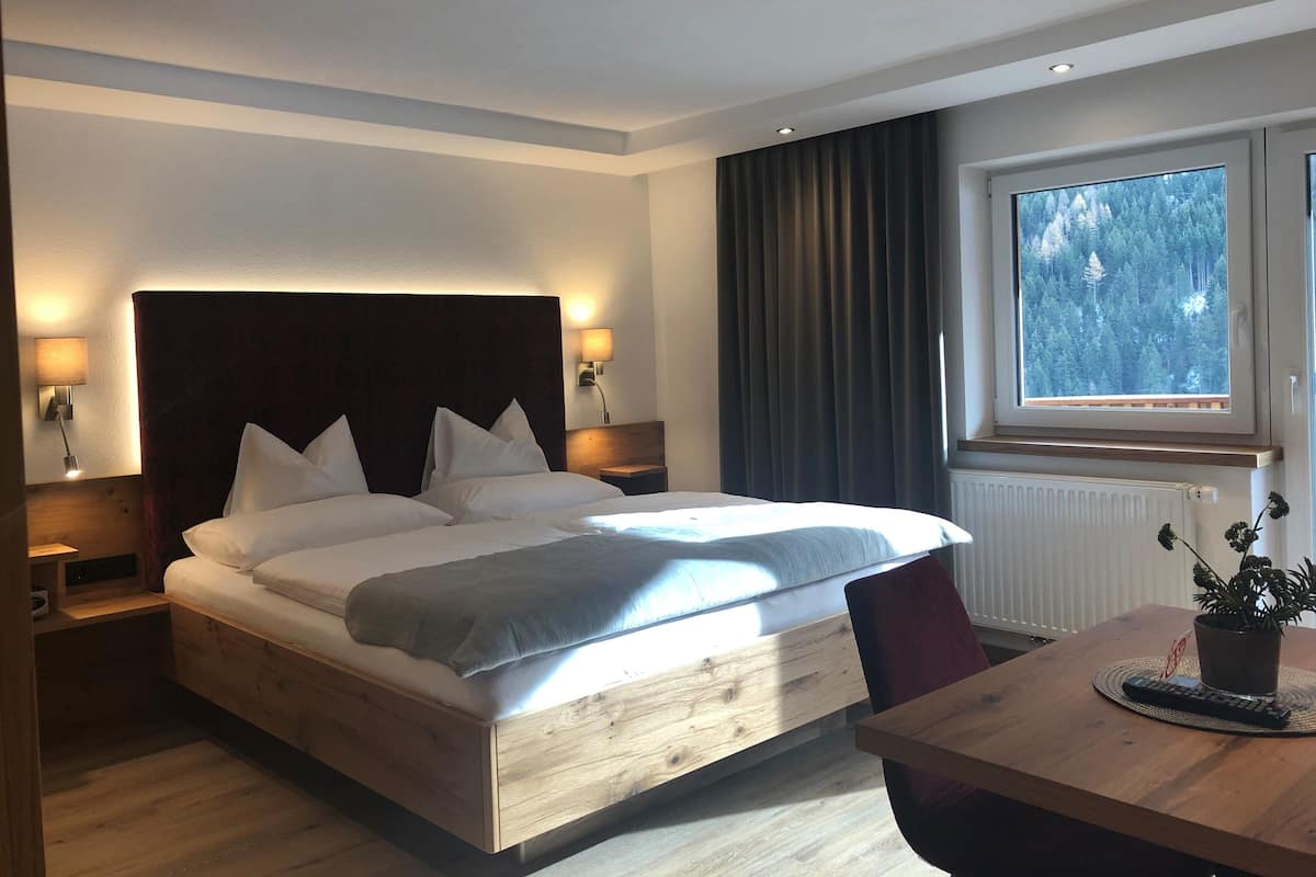 A modern hotel room with a large bed, wooden headboard, two bedside lamps, a window with a forest view, and a wooden table with a remote control.