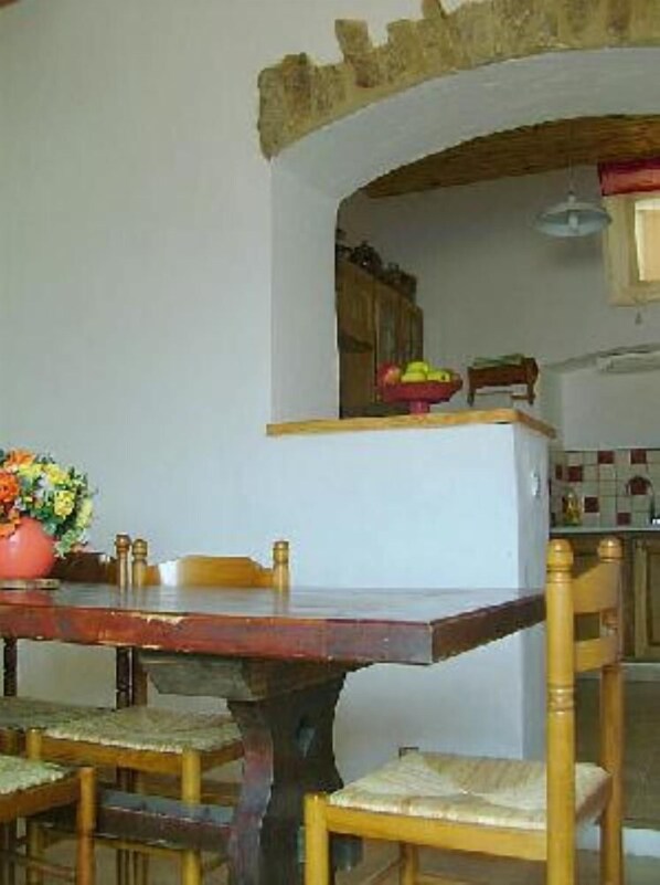 Una mesa de comedor con sillas, un jarrón de flores y una zona de cocina con armarios y un frutero.