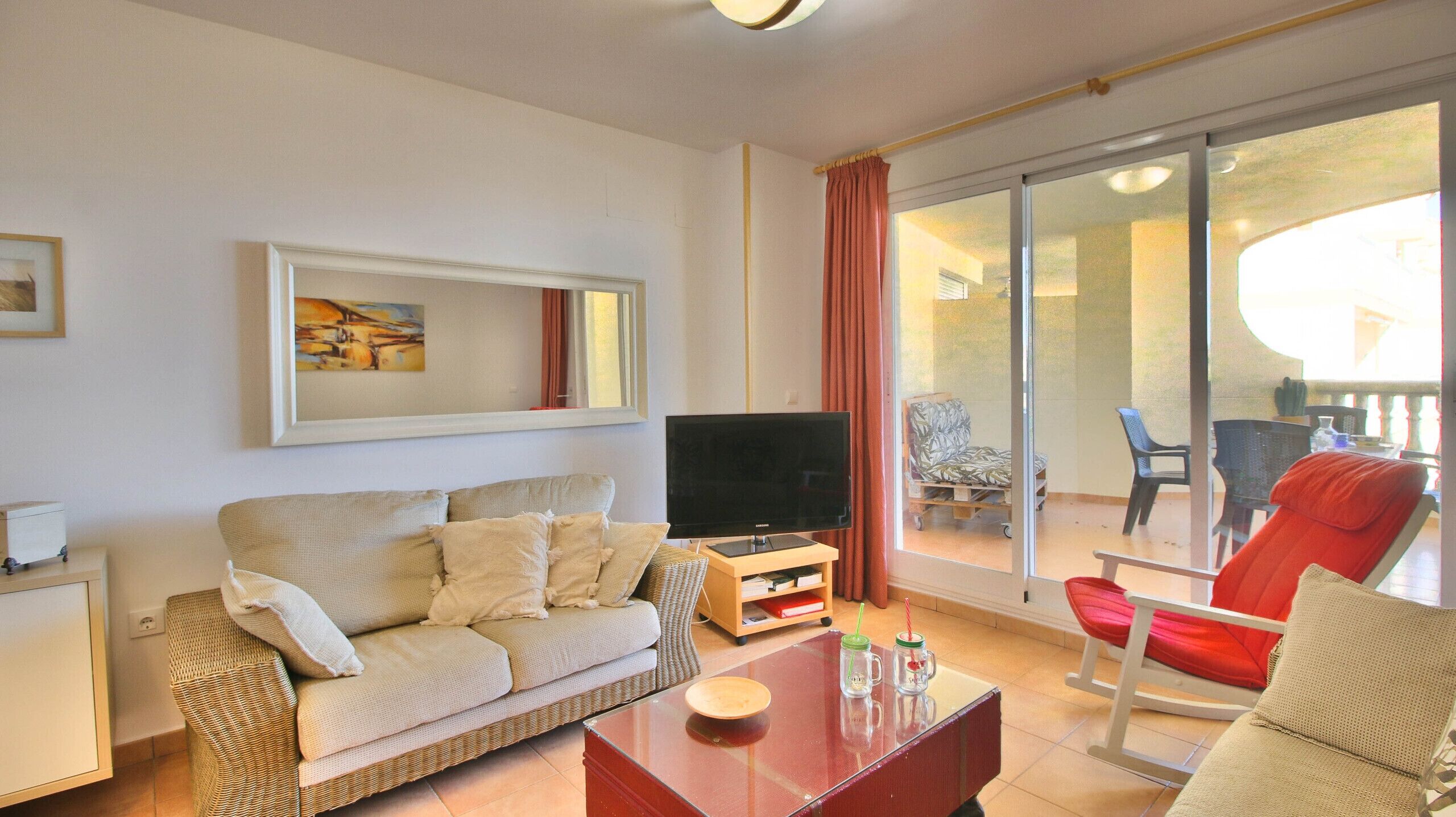 A living room with a sofa, a coffee table, a TV, and a balcony with chairs.