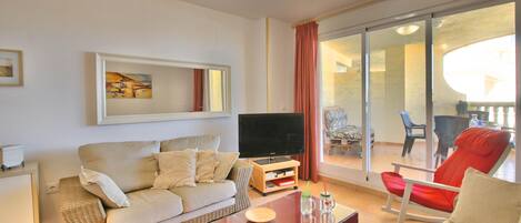 A living room with a sofa, a coffee table, a TV, and a balcony with chairs.