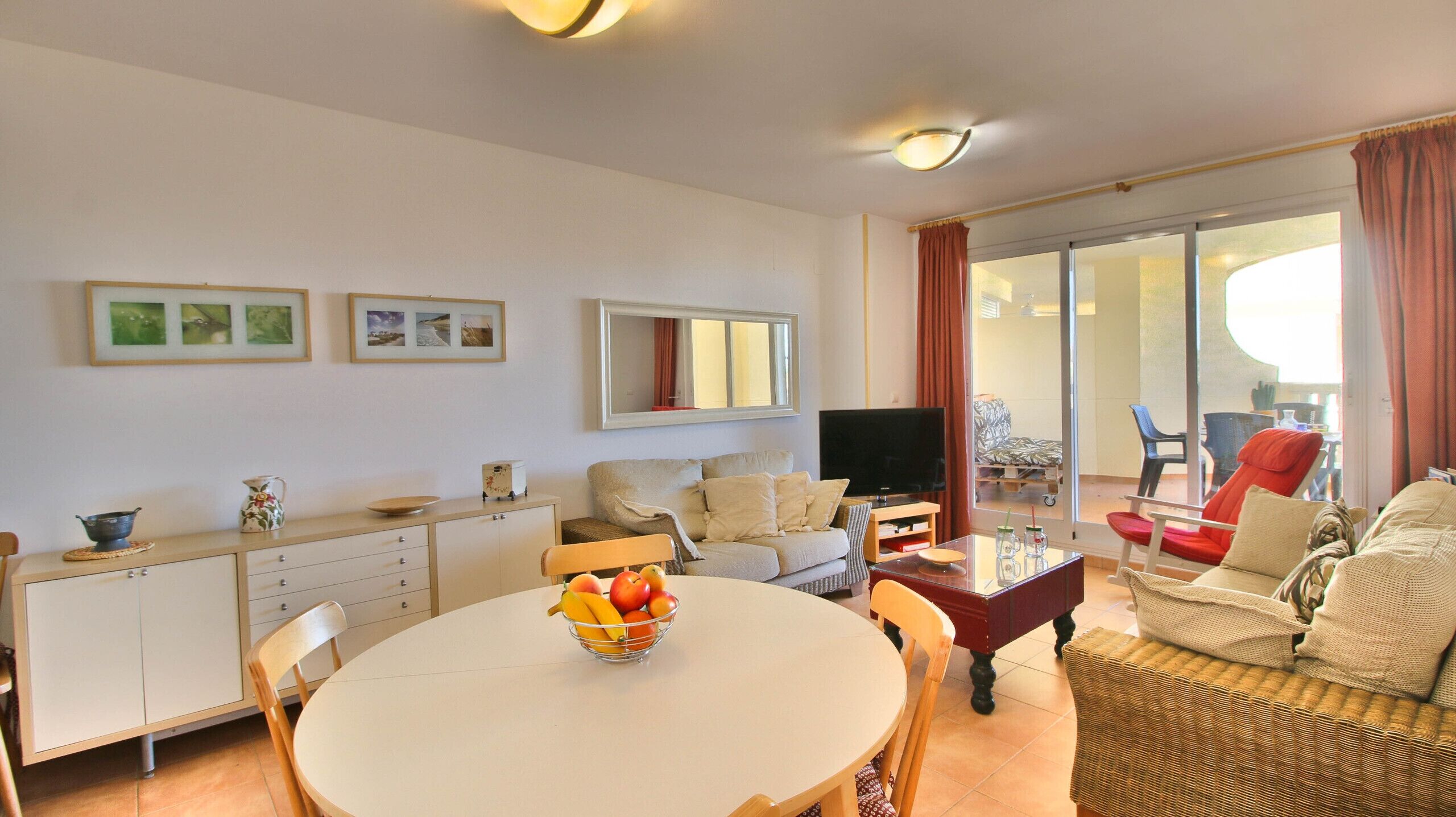 Una sala de estar con un sofá, un televisor, una mesa de comedor con fruta y un balcón con una silla.
