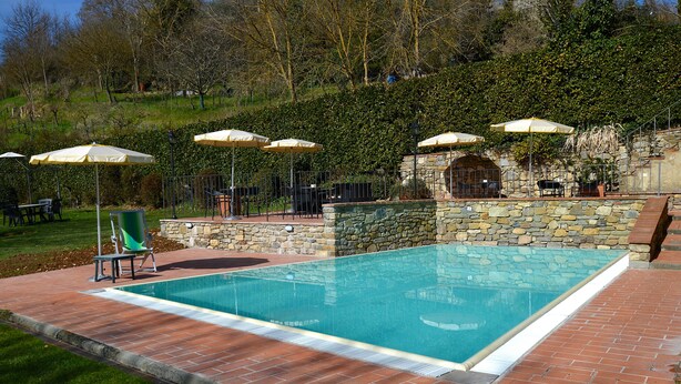 Ein rechteckiger Swimmingpool mit einer Steinmauer und einer gefliesten Liegefläche. Draussen stehen Stühle und Sonnenschirme.