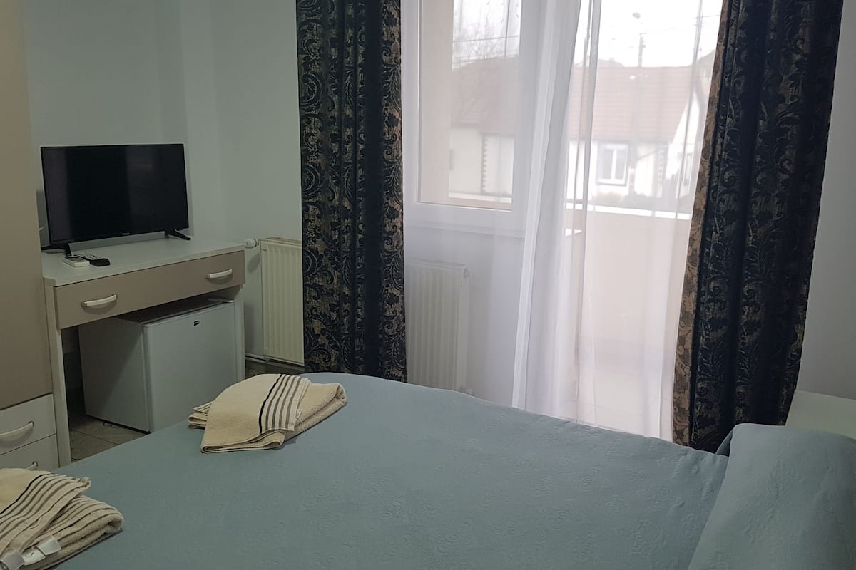 A hotel room with a bed, a desk with a television, a small refrigerator, and curtains.