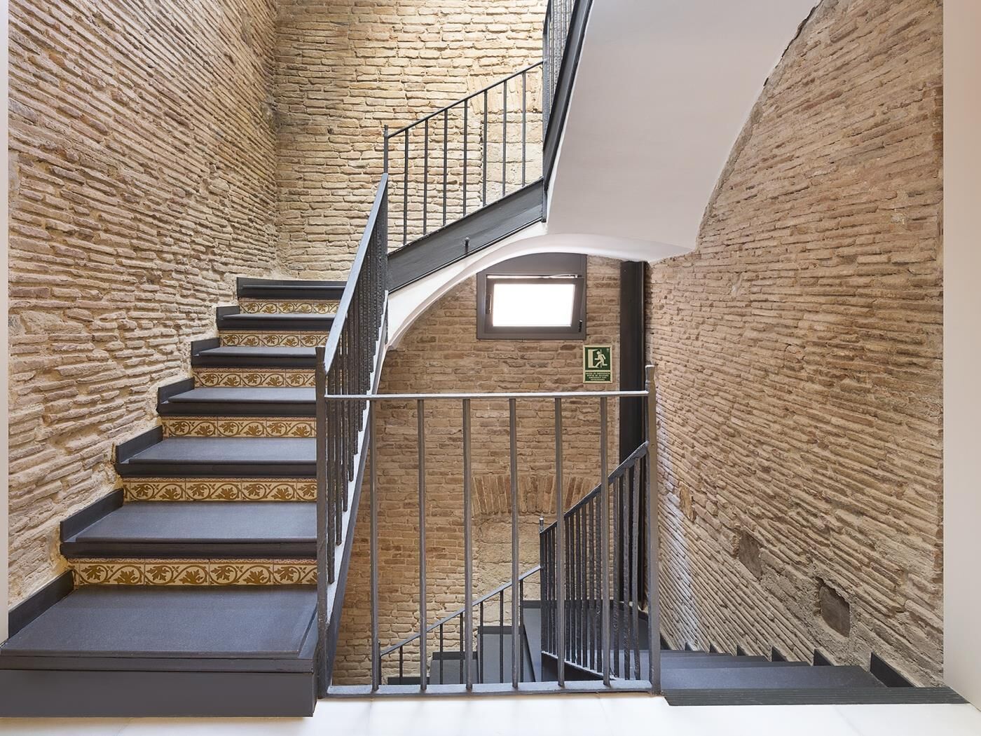 Un escalier, avec un mur de briques d’un côté et un mur blanc de l’autre, qui mène à l’étage supérieur.