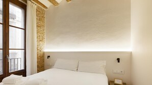 A bedroom with a large bed, white linens, a wooden headboard, a brick wall, and a window with curtains. - Deluxe One Bedroom Apartment (Barcelona)