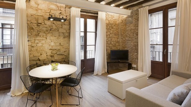 A cozy living room with a stone wall, a round dining table with chairs, a flat-screen TV, and a sofa. - Deluxe One Bedroom Apartment (Barcelona)