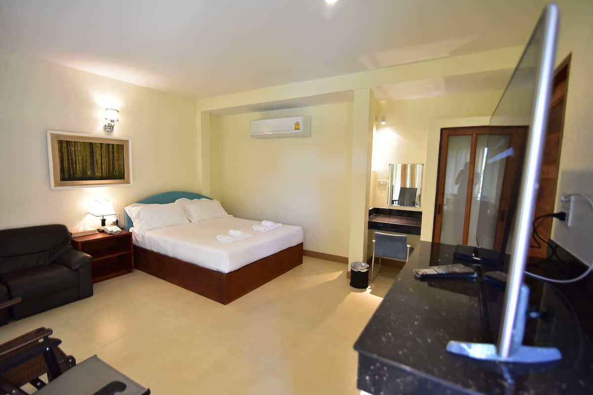 A hotel room with a bed, a desk with a computer, a chair, a lamp, a trash bin, and an air conditioning unit.