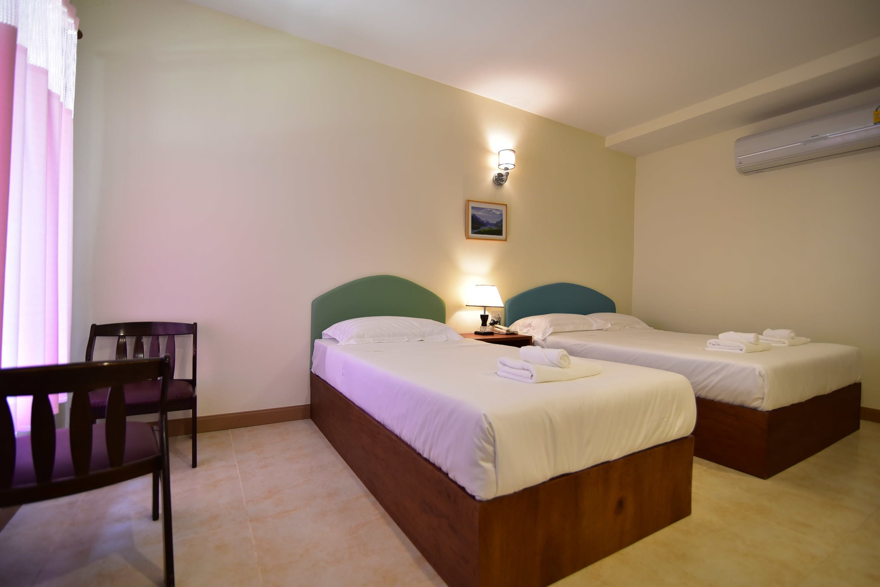 A hotel room with two single beds, a chair, a nightstand with a lamp, and an air conditioning unit on the wall.