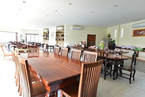 Uma sala de jantar ampla com várias mesas e cadeiras de madeira, um aparelho de ar condicionado na parede e uma porta que dá para outro quarto.