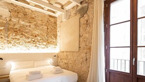 Une chambre avec un mur en pierre, un lit recouvert de draps blancs, une porte en bois et une fenêtre avec des rideaux.
