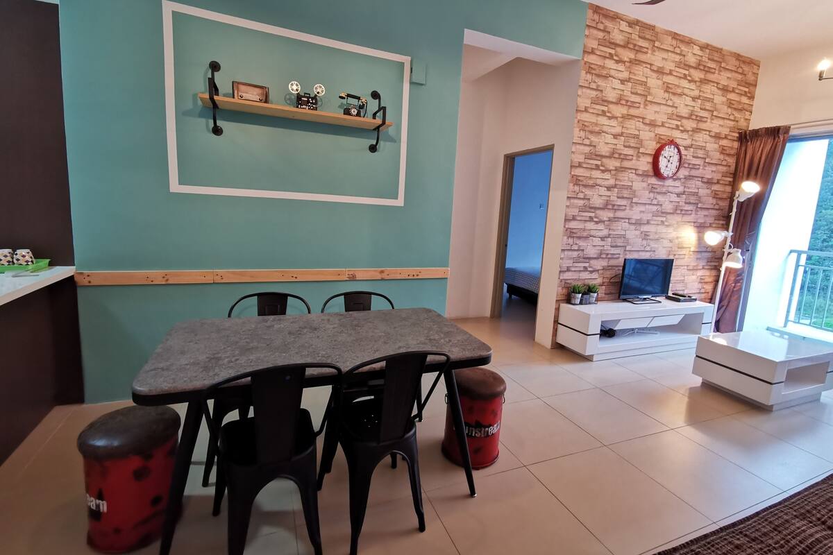 A dining area with a table and chairs, a wooden shelf with decorative items, a brick accent wall, a TV, and a clock.