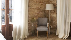 Een hoekappartement met een bakstenen muur, een houten vloer, een grijze fauteuil, een vloerlamp en een raam met gordijnen.
