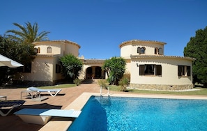 A pool area with lounge chairs and a clear blue pool, surrounded by a building with a tiled roof and stone base.