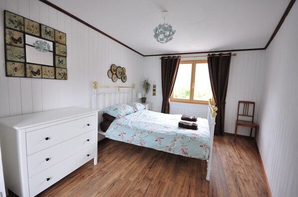 Un dormitorio con piso de madera, un tocador blanco con cajones, una cama con ropa de cama floral, un candelabro, una ventana con cortinas y una silla de madera.