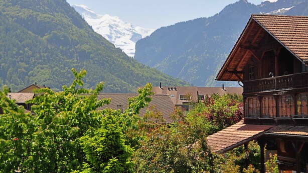 A wooden chalet with a sloped roof, surrounded by lush greenery and mountains in the background. - Beautifully designed Apartment.  Fully equipped for your holiday needs. Sleeps 7 (Interlaken)