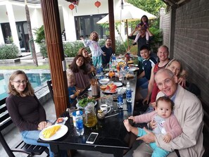Un grupo de personas sentadas alrededor de una mesa con comida y bebidas, al aire libre, con una piscina y vegetación de fondo.