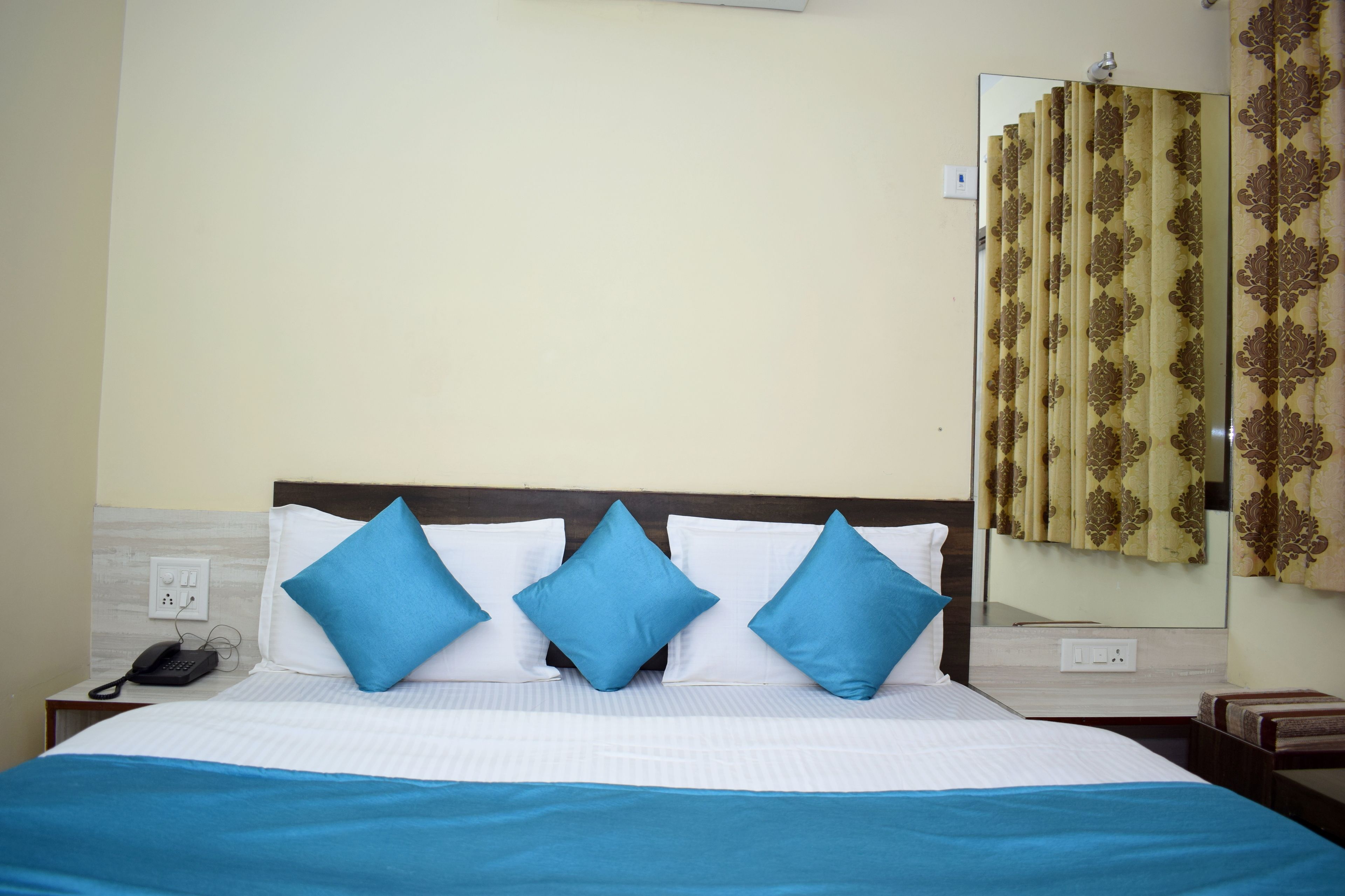 A neatly made bed with blue pillows and a white bedspread, a bedside phone, and a mirror reflecting a patterned curtain.
