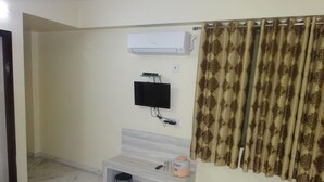 Una habitación de hotel con cama, una mesita, un banco, un equipo de aire acondicionado, un televisor y una cortina.