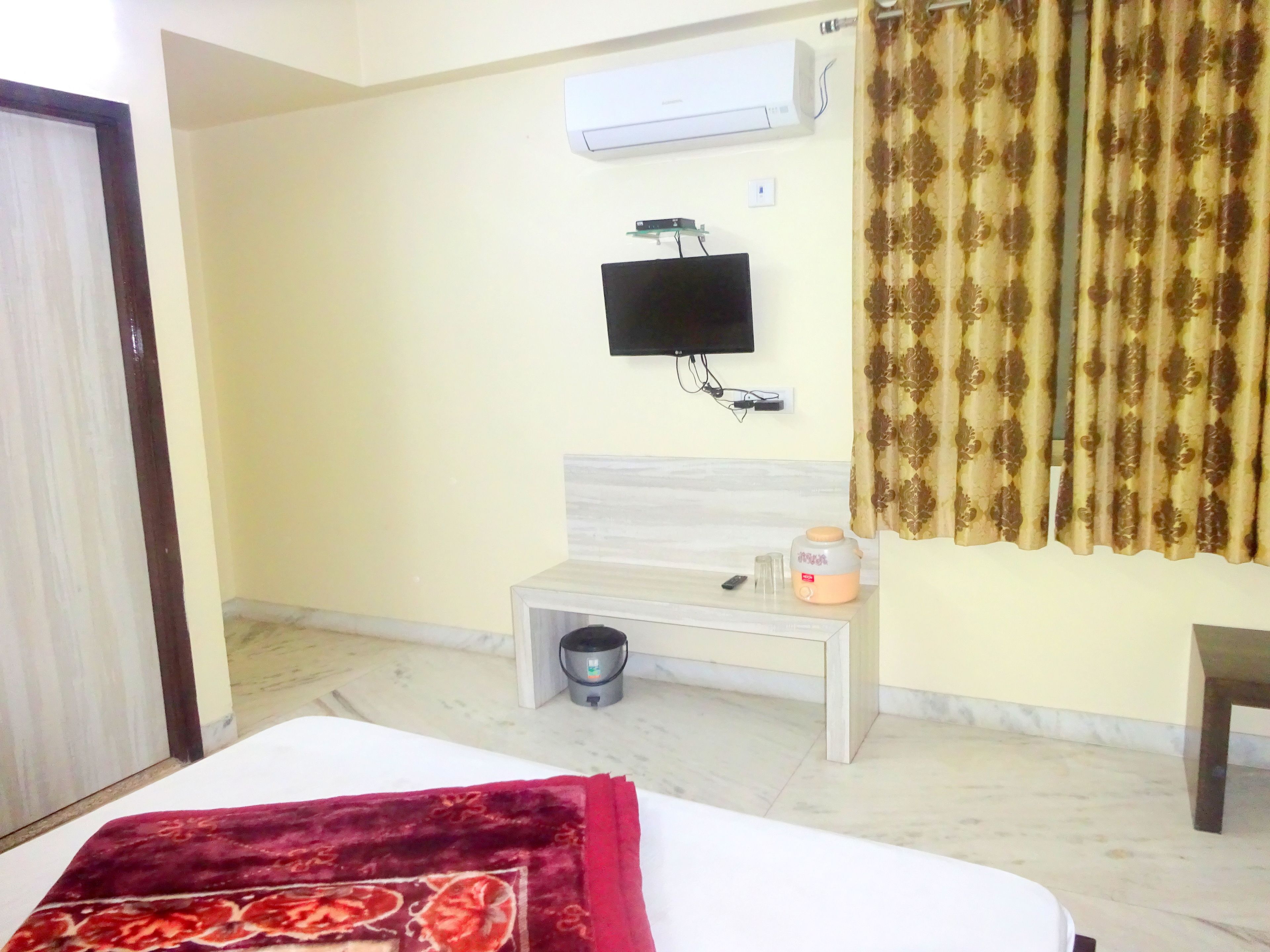 Habitación de hotel con cama, televisión en la pared, una mesita con un control remoto y una cortina.