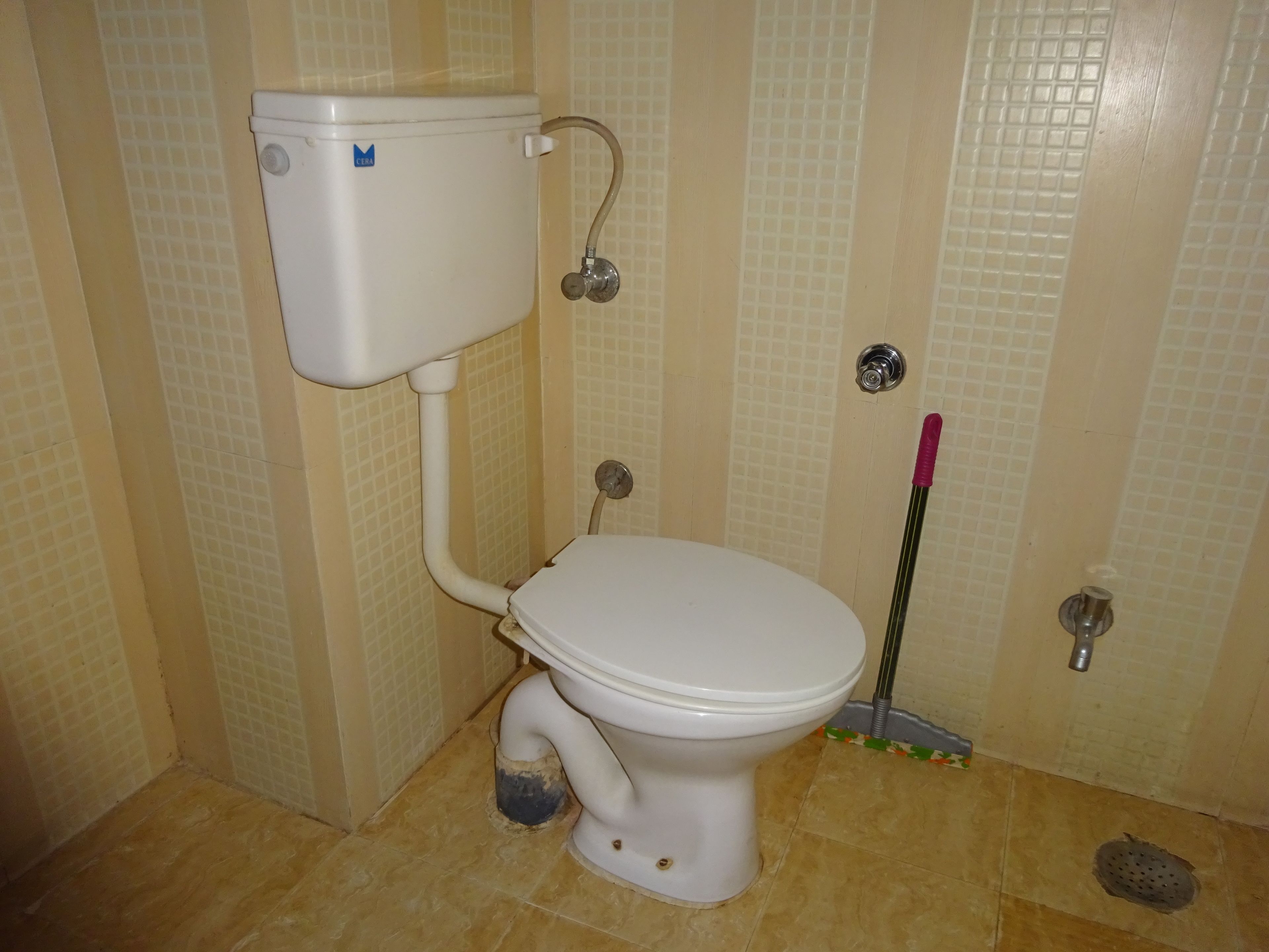 A toilet with a white tank and seat, a visible pipe, and a mop leaning against the wall.