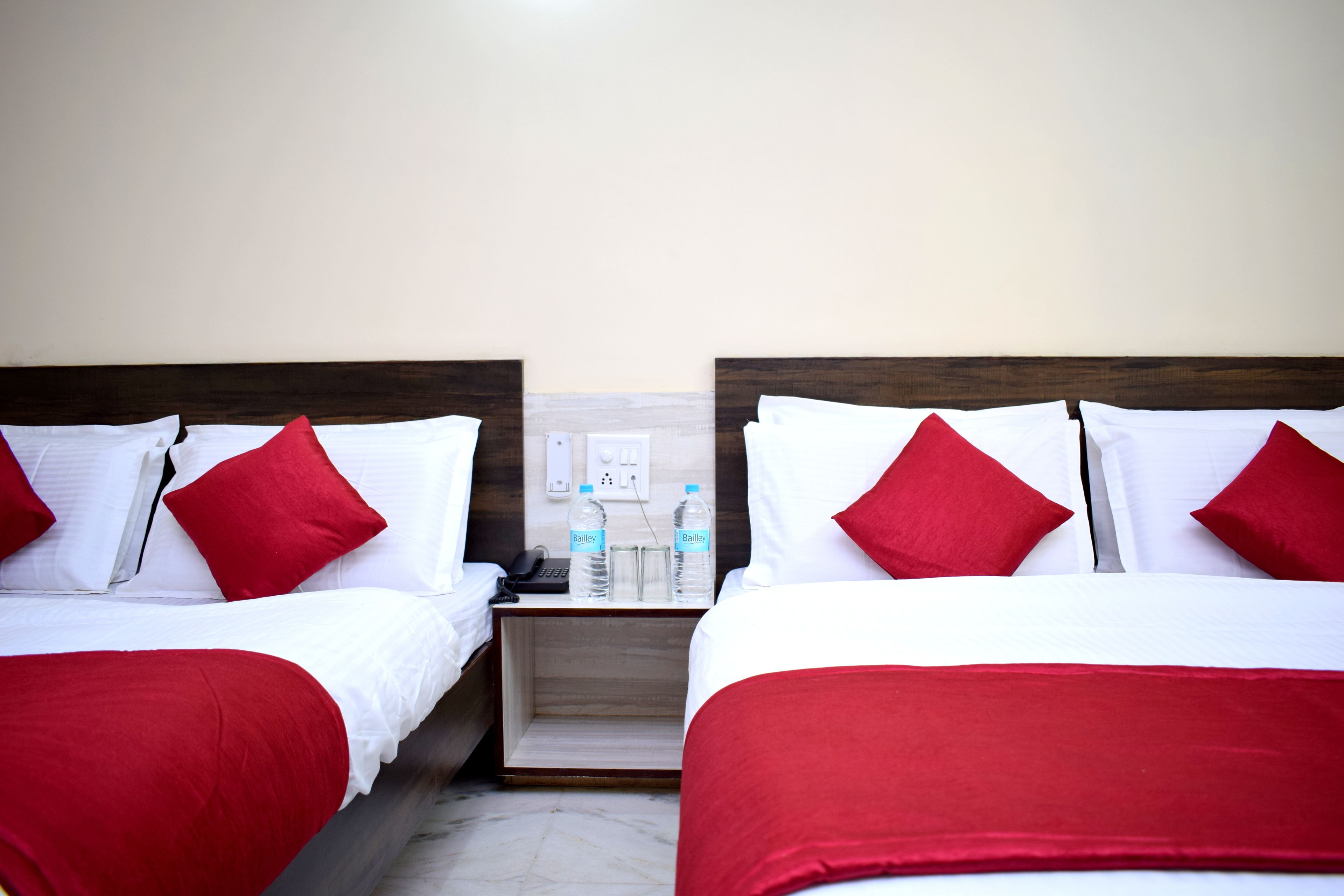 A hotel room with two beds, each with white bedding and red pillows. There is a nightstand with two water bottles and a phone between the beds.