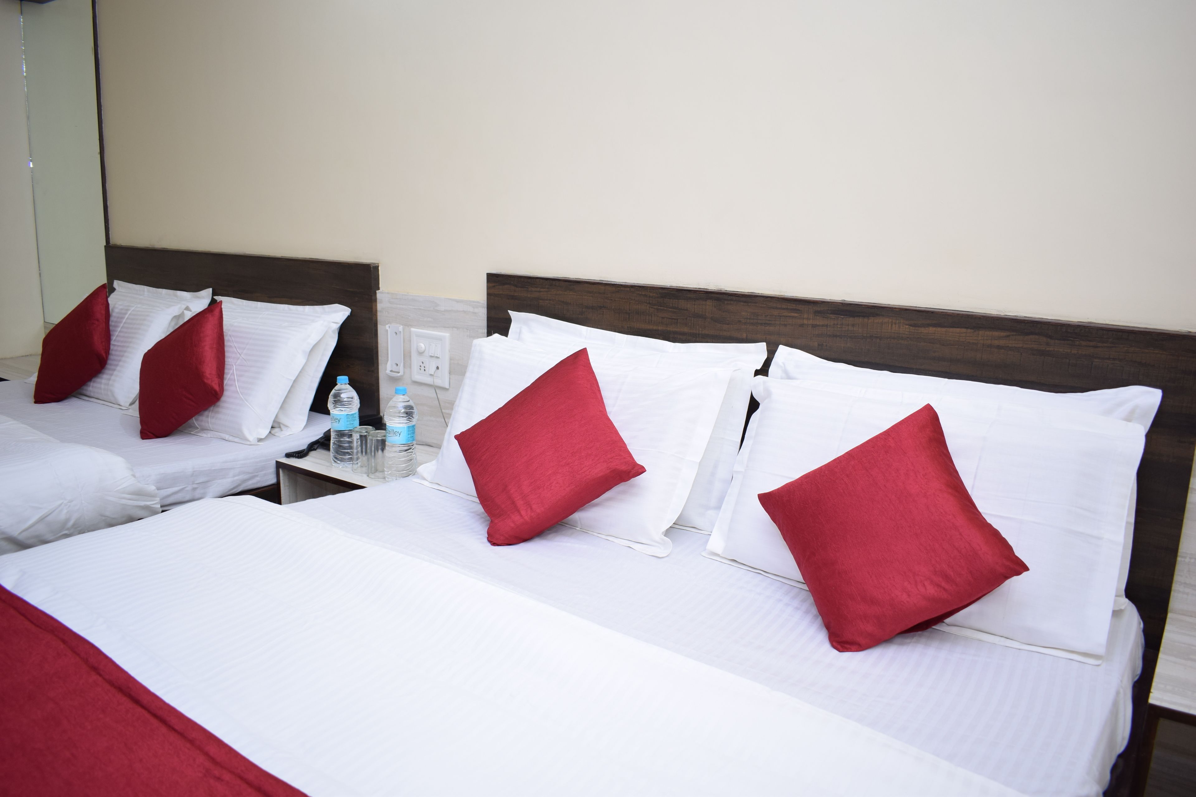 A hotel room with two beds, each with white bedding and red pillows. There is a nightstand with a water bottle and glasses.
