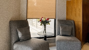 A modern interior with two grey upholstered chairs, a round table with a vase of flowers, and a large window with a beige blind.