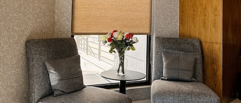 Un interior moderno con dos sillones tapizados grises, una mesa redonda con un jarrón de flores y un ventanal grande con persiana color beige.