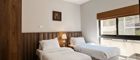 A modern hotel room with two beds, a wooden wardrobe, a small table, and a window with a view of the outside.