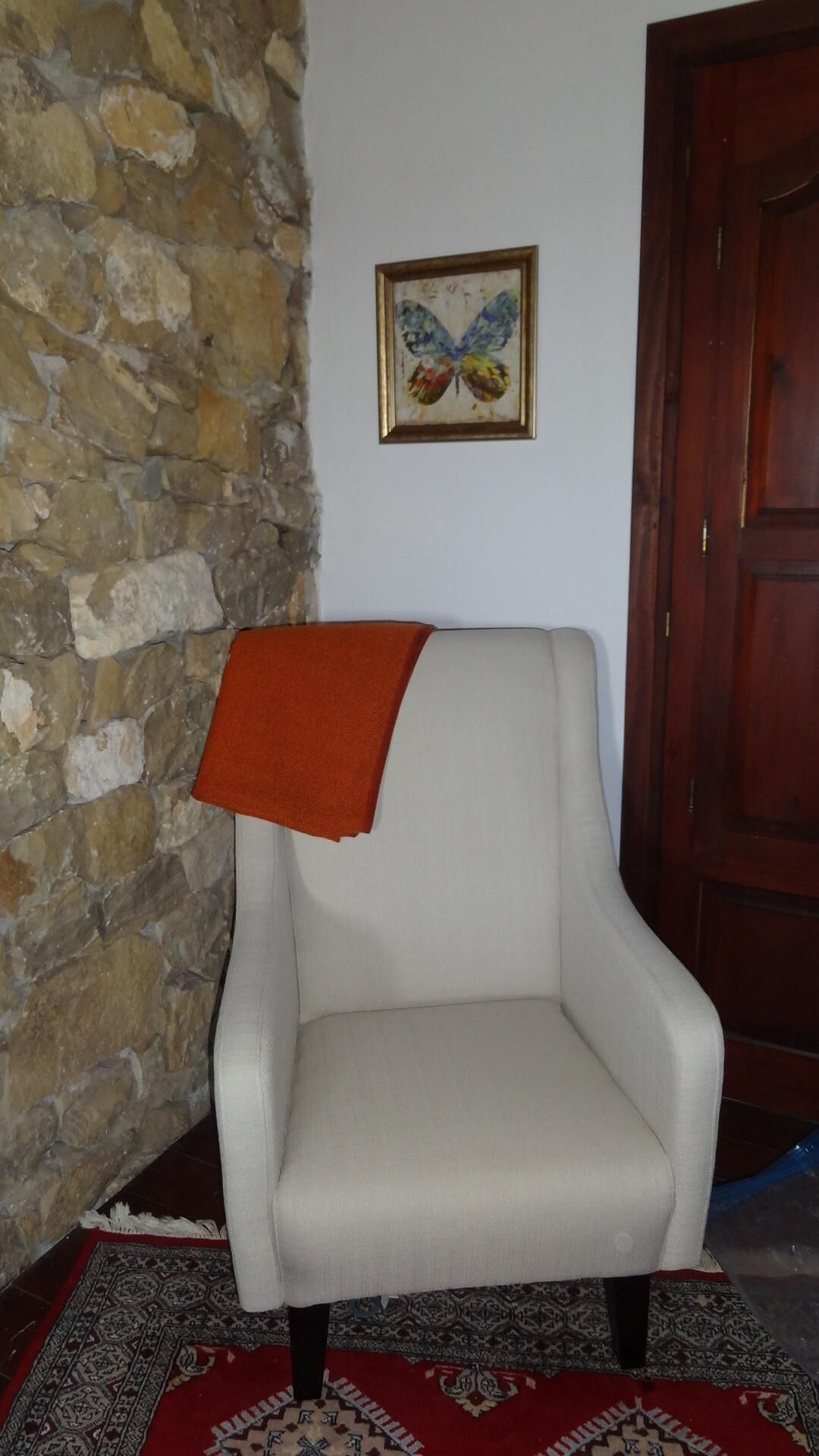 Ein Zimmer mit einer Steinsäule, einem weißen Sessel mit roter Decke, einer Holztür und einem Bilderrahmen mit einem Schmetterlingsgemälde an der Wand.