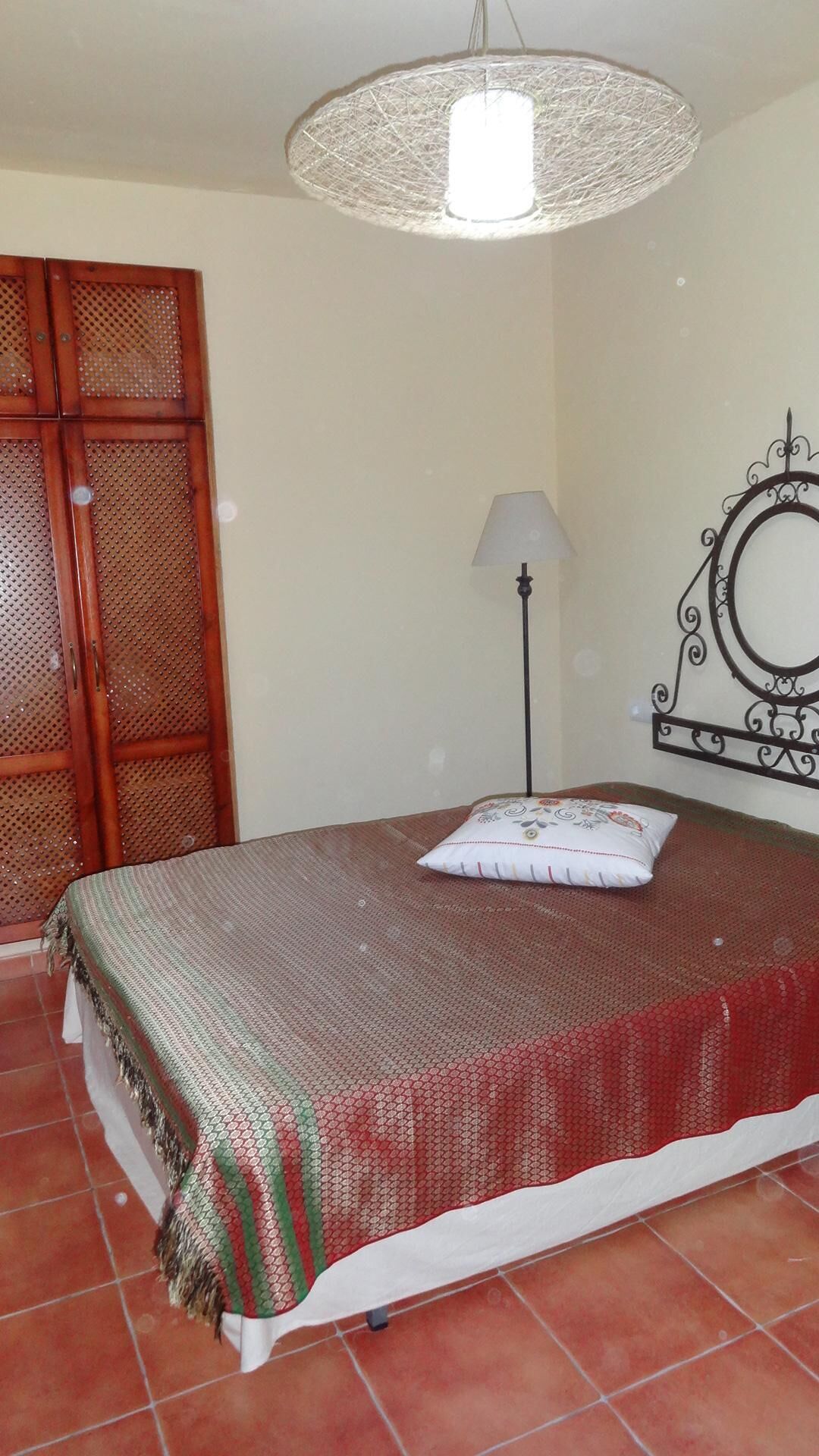 Een slaapkamer met een bed, een hanglamp, een vloerlamp, een kledingkast en een decoratieve spiegel.