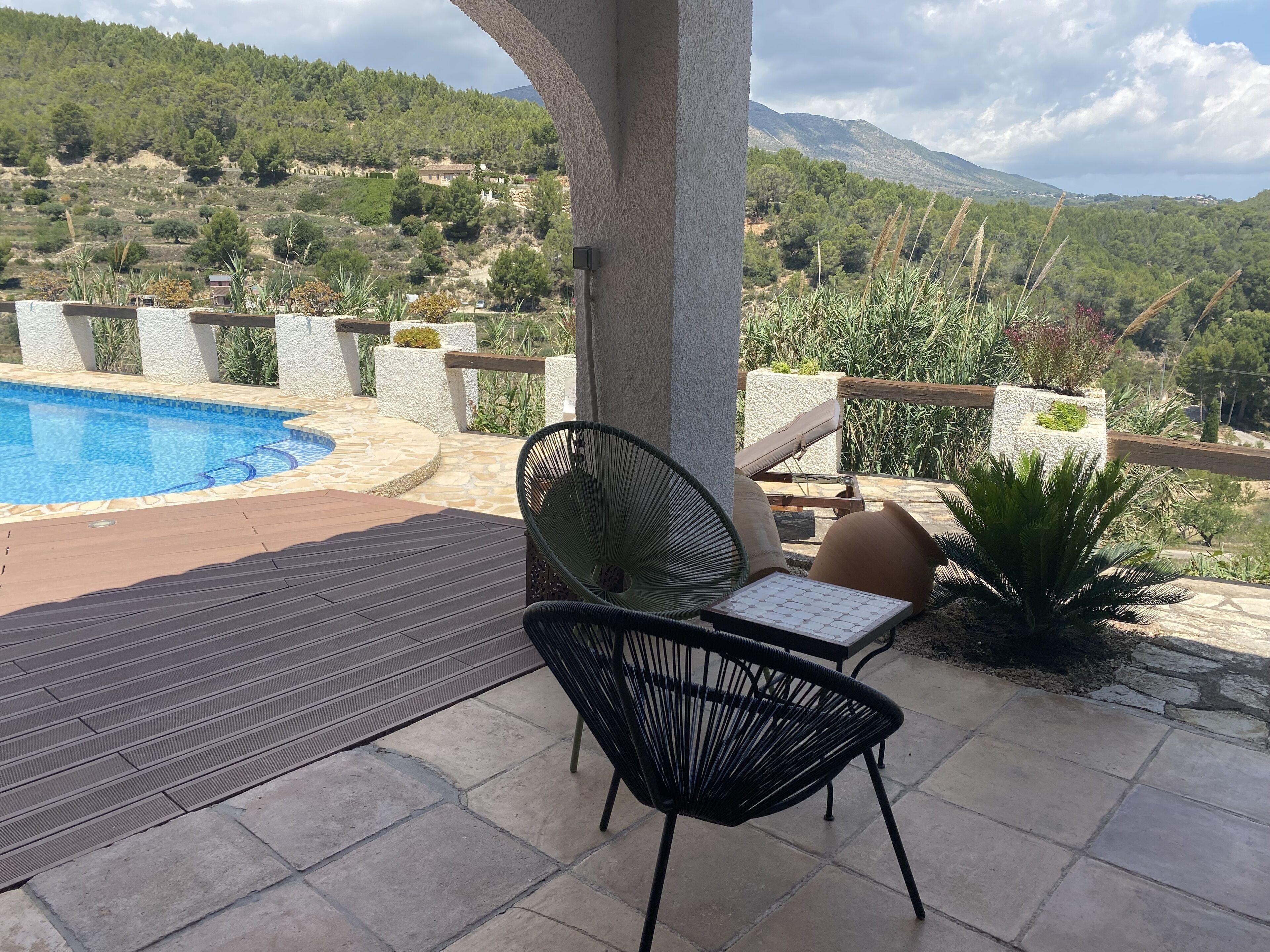 A poolside area with a chair and a table, surrounded by stone flooring and a view of a mountainous landscape.