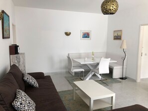 A modern living room with a brown sofa, a white coffee table, and a dining area with a white table and chairs. There is a decorative lamp and a framed picture on the wall.
