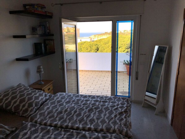 A bedroom with a bed, a nightstand, a lamp, a shelf with books, and a sliding glass door leading to a balcony with a view of greenery and a building.