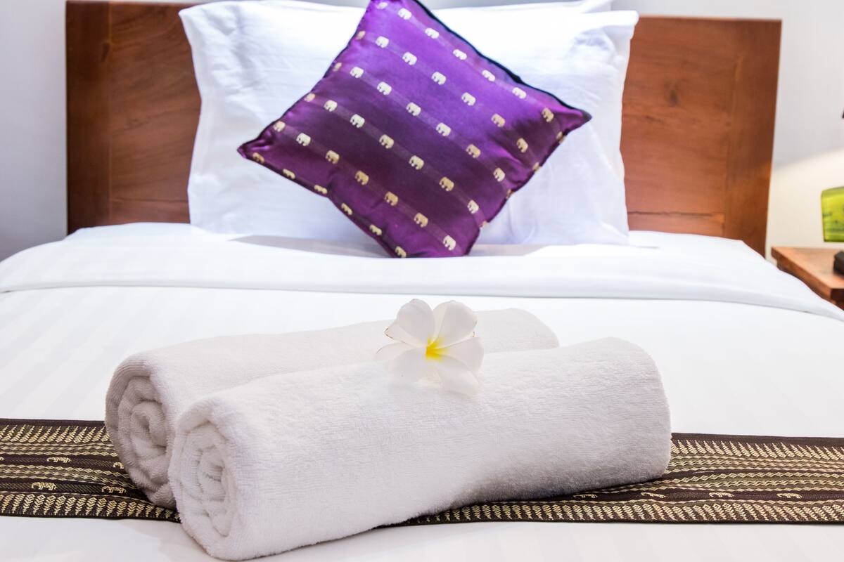 A neatly made bed with a white towel, a purple pillow with a pattern, and a white flower.