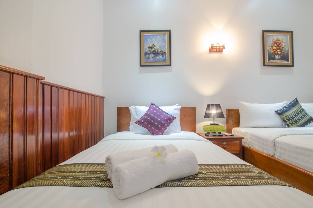 A hotel room with two beds, wooden headboards, white bedding, a green and gold patterned bedspread, a purple pillow, a white towel with a flower, a nightstand with a lamp, and two framed pictures on the wall.