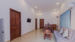 A living room with a sofa, a dining table, and a TV. There are two wooden doors and a ceiling fan. - Aster Villa (Siem Reap)
