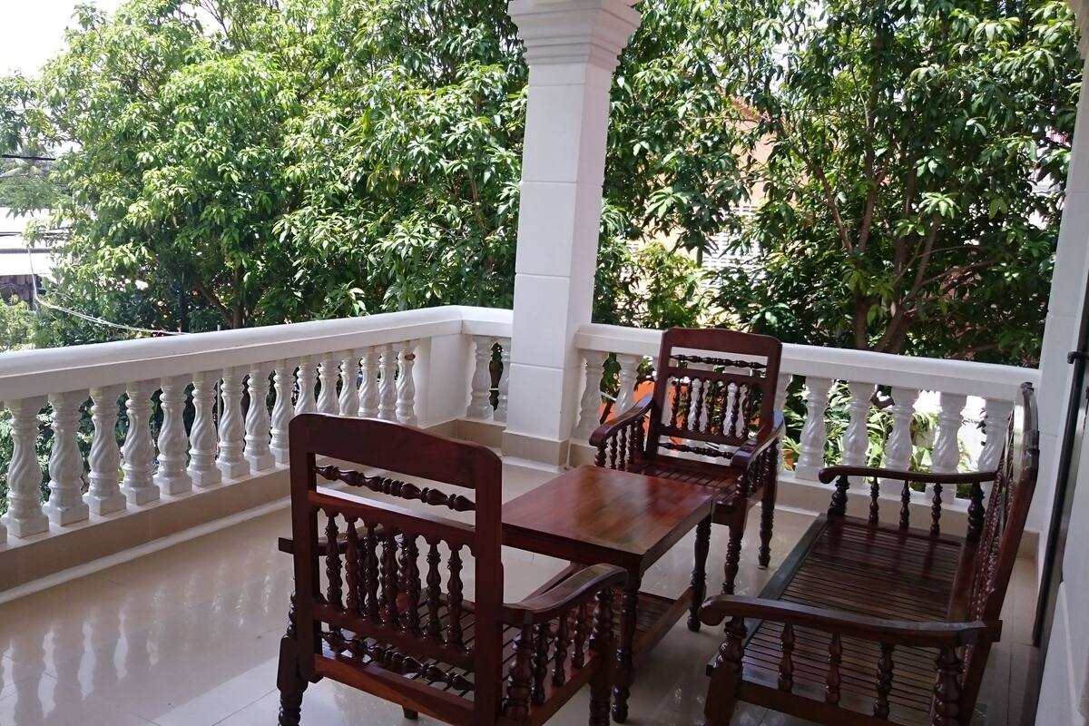 A balcony with a wooden table and chairs, white balustrades, and lush greenery in the background.