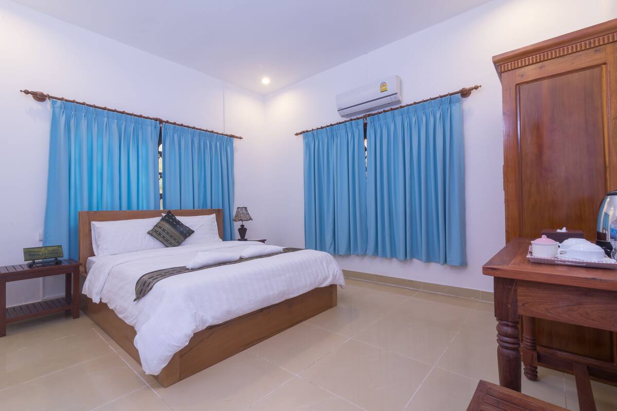 A bedroom with a wooden bed, blue curtains, a wooden nightstand, a table with a tray, and an air conditioning unit.