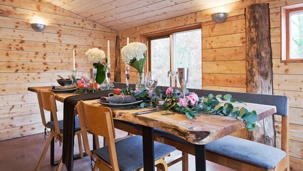 A wooden cabin interior with a dining table set for four, surrounded by chairs, and a large window providing natural light. - The elbowroom escape (Donard)