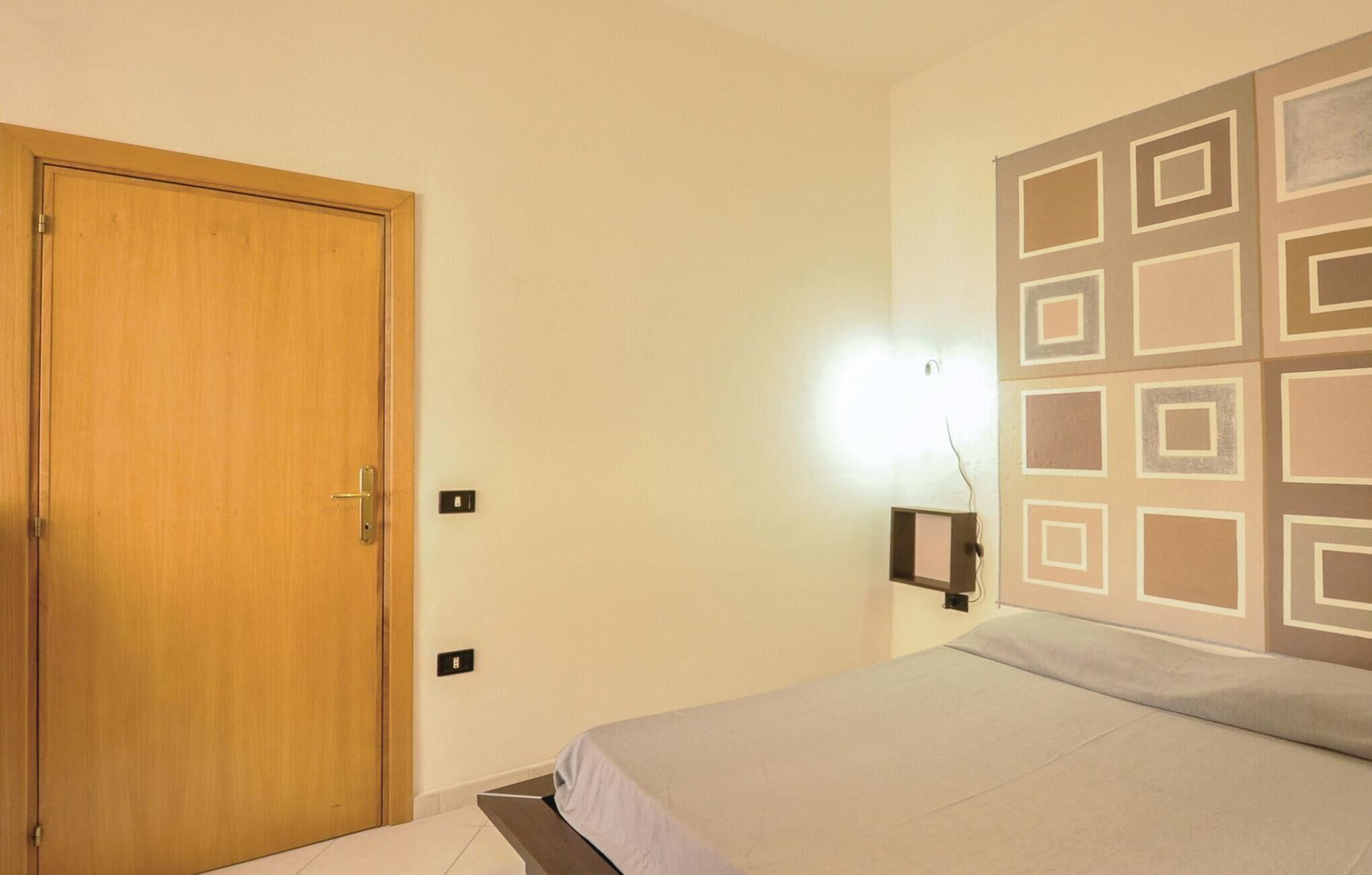 A bedroom with a wooden door, a bed with a light-colored bedspread, a wall-mounted light, and a decorative wall panel.